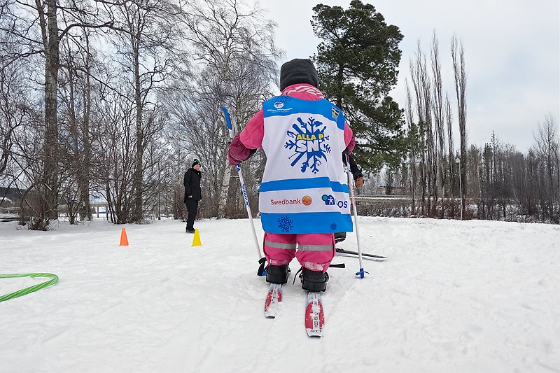 Ett litet barn i rosa overall och Alla på snö-väst testar längdskidor.